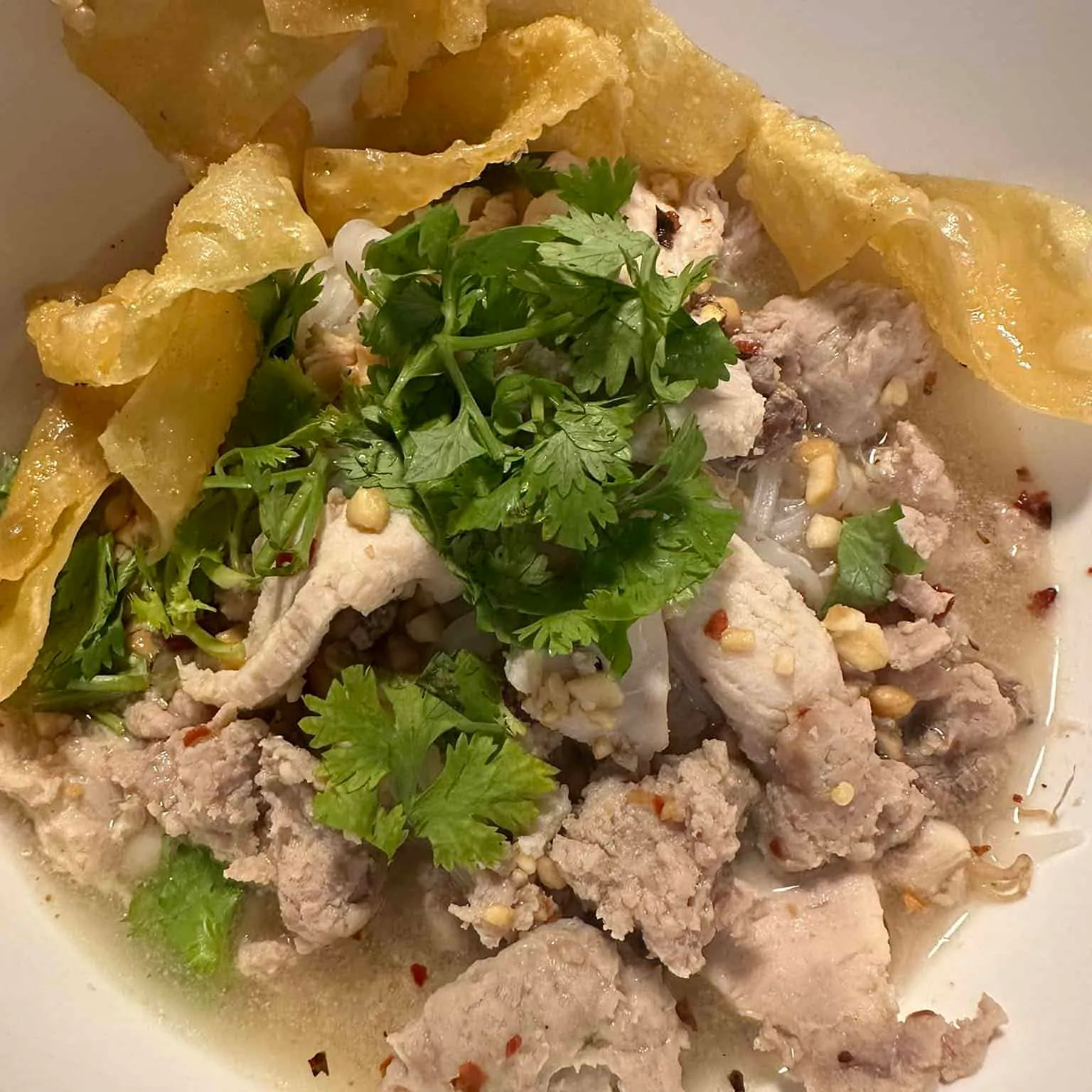 Lys suppe med strimlet svinekjøtt, koriander og friterte chips i dyp skål.